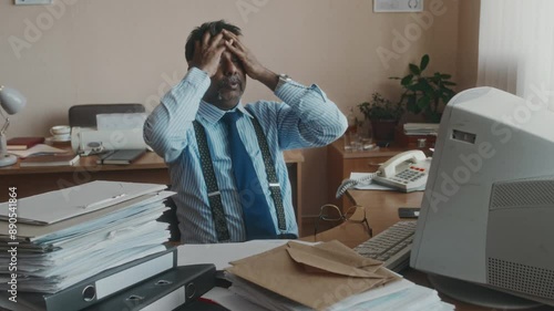 Medium shot of mature Biracial employee in formalwear working with myriads of documents, taking off glasses and putting arms around head, having severe headache from monotonous work in retro office