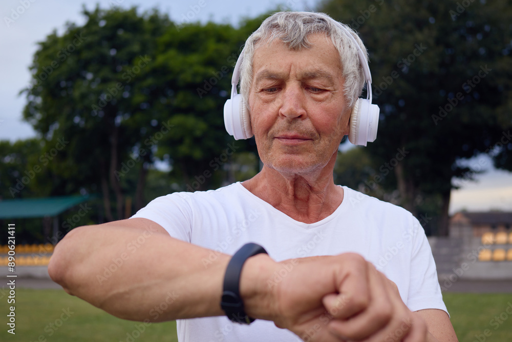Middle-aged man in a white t-shirt checking his smartwatch for heart rate data standing at the stadium wearing wireless headphones using modern technology for sports workouts outdoor Middle-aged man in a white t-shirt checking his smartwatch for heart rate data standing at the stadium wearing wireless headphones using modern technology for sports workouts outdoor