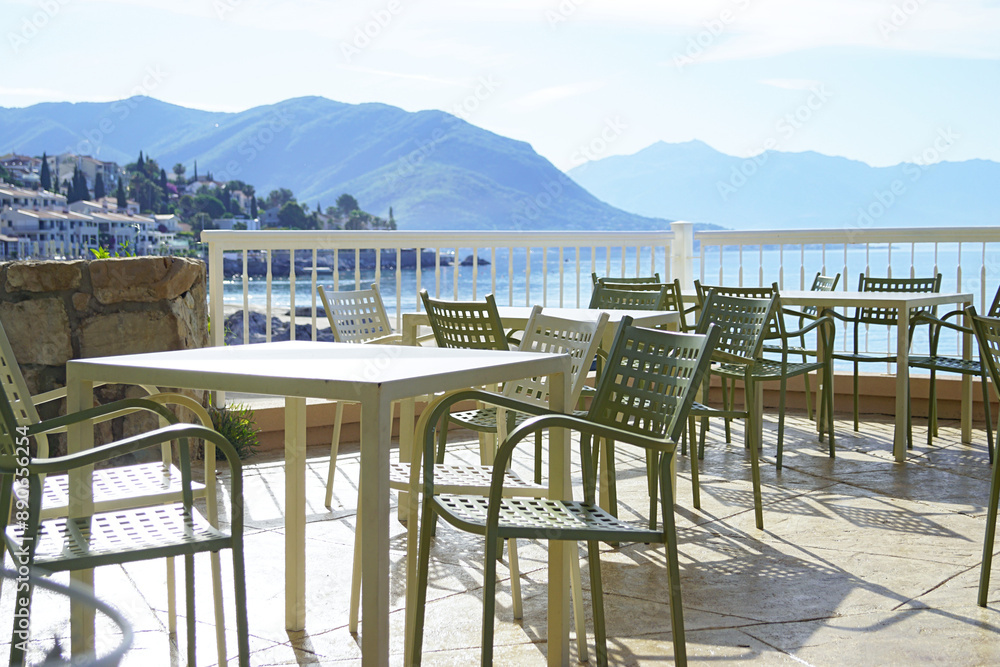 Early sunny morning on the embankment of the city of Herceg Novi, Montenegro: empty tables and chairs on the terrace of a cafe against the backdrop of the city, sea and mountains. Early sunny morning on the embankment of the city of Herceg Novi, Montenegro: empty tables and chairs on the terrace of a cafe against the backdrop of the city, sea and mountains.