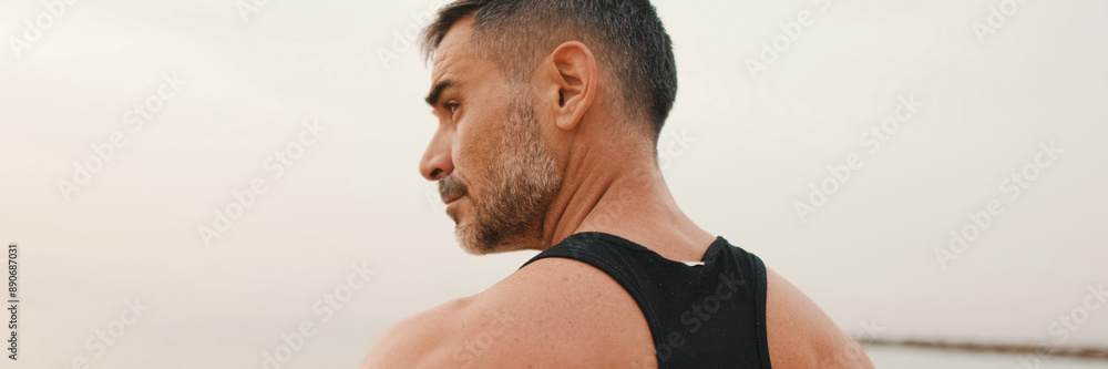 Obraz premium Close-up of middle-aged athletic man standing on the beach after workout and looking at the seascape, panorama
