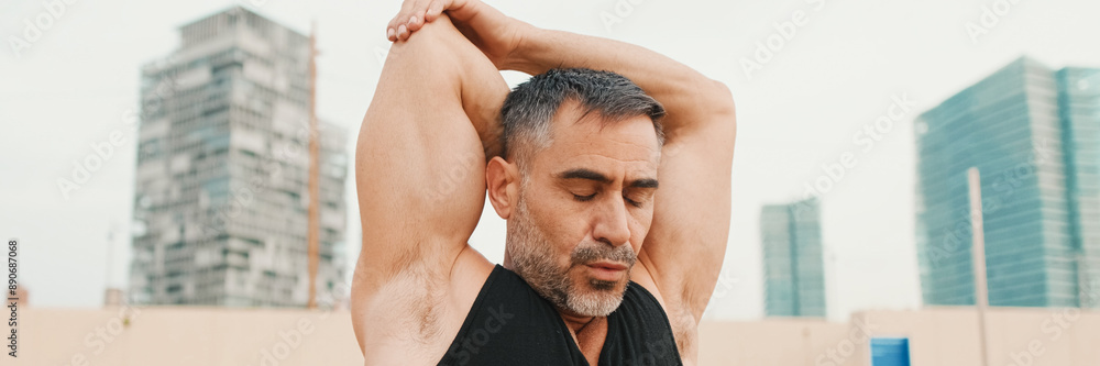Fototapeta premium Athletic mature man stretches arm muscles while standing on the beach in the morning