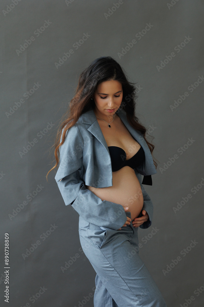 A beautiful pregnant brunette woman in a cropped gray stylish suit on a gray paper background in the studio. A pretty girl with a bare stomach in a fashionable suit is sitting barefoot on a gray backg A beautiful pregnant brunette woman in a cropped gray stylish suit on a gray paper background in the studio. A pretty girl with a bare stomach in a fashionable suit is sitting barefoot on a gray backg
