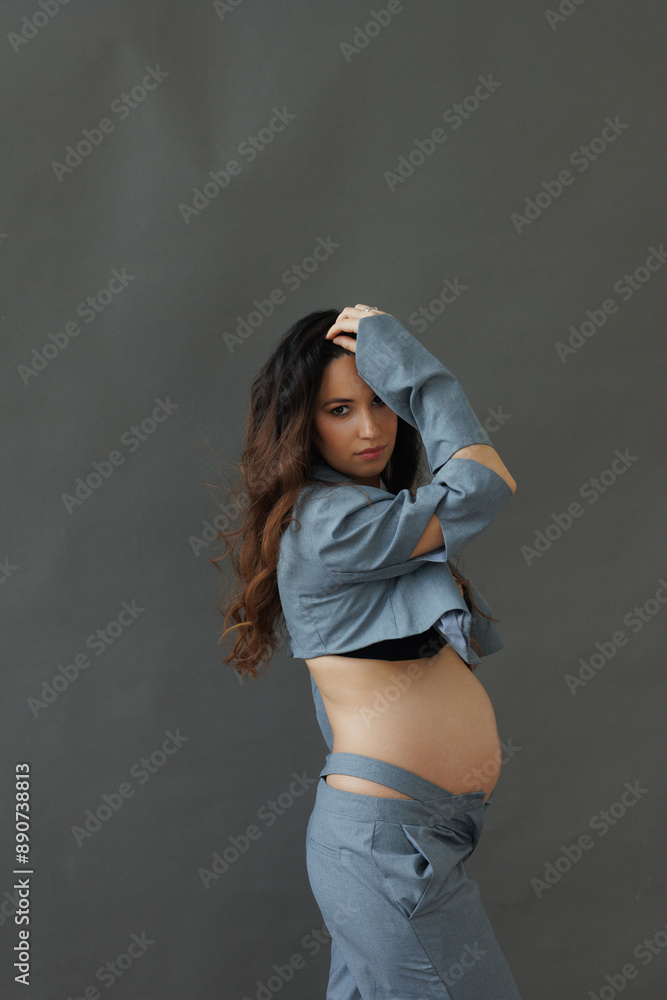 A beautiful pregnant brunette woman in a cropped gray stylish suit on a gray paper background in the studio. A pretty girl with a bare stomach in a fashionable suit is sitting barefoot on a gray backg
