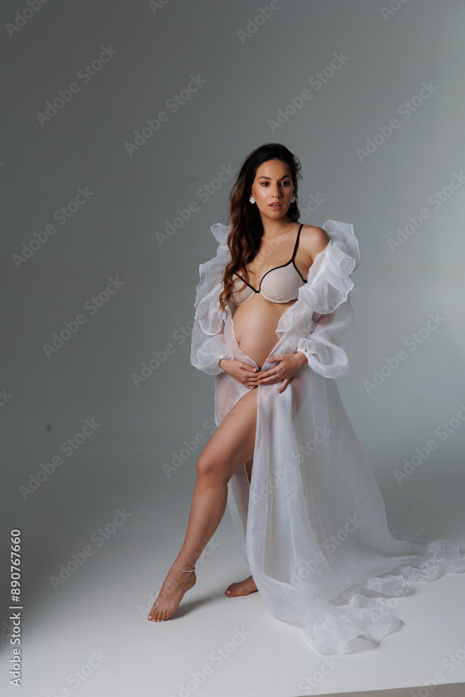 A beautiful pregnant brunette woman in a white flying transparent negligee, dressing gown. A pretty girl with a bare stomach in lingerie stay barefoot on a grey background. Smile, portrait A beautiful pregnant brunette woman in a white flying transparent negligee, dressing gown. A pretty girl with a bare stomach in lingerie stay barefoot on a grey background. Smile, portrait