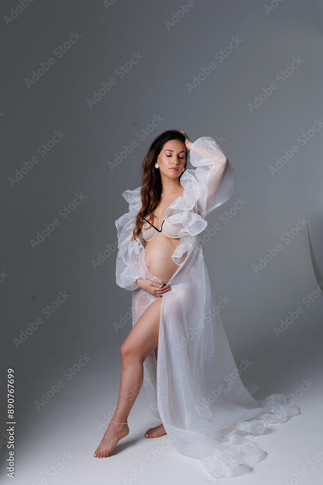 A beautiful pregnant brunette woman in a white flying transparent negligee, dressing gown. A pretty girl with a bare stomach in lingerie stay barefoot on a grey background. Smile, portrait A beautiful pregnant brunette woman in a white flying transparent negligee, dressing gown. A pretty girl with a bare stomach in lingerie stay barefoot on a grey background. Smile, portrait