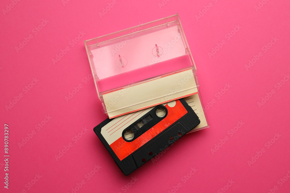 A cassette tape is laying on a pink background. The tape is red and black