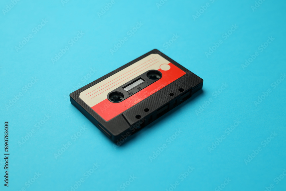 A black and red cassette tape sits on a blue surface. The tape is old and worn, with a red stripe running down the middle. The tape appears to be a vintage item, possibly from the 1980s or 1990s A black and red cassette tape sits on a blue surface. The tape is old and worn, with a red stripe running down the middle. The tape appears to be a vintage item, possibly from the 1980s or 1990s
