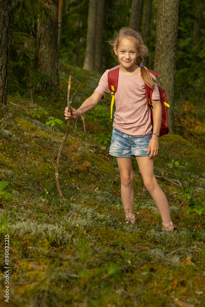 A young girl wearing a pink shirt, floral denim shorts, and a red backpack holds a stick while hiking through a forest, smiling and enjoying the adventure. A young girl wearing a pink shirt, floral denim shorts, and a red backpack holds a stick while hiking through a forest, smiling and enjoying the adventure.