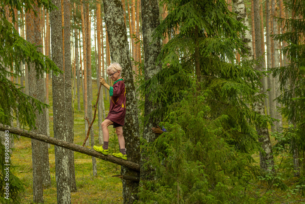 A young boy in a maroon shirt and yellow-green shoes climbs a tree in a dense forest, holding a stick and looking at the camera. A young boy in a maroon shirt and yellow-green shoes climbs a tree in a dense forest, holding a stick and looking at the camera.