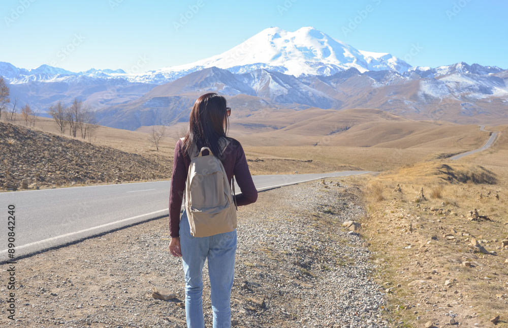 A female traveler with a backpack walks along a road in the mountains. View of Elbrus, North Caucasus, Russia