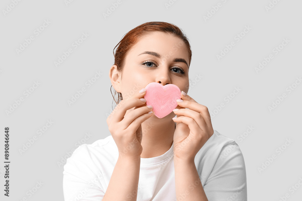 Young woman covering mouth with heart-shaped silicone facial cleansing brush on white background Young woman covering mouth with heart-shaped silicone facial cleansing brush on white background