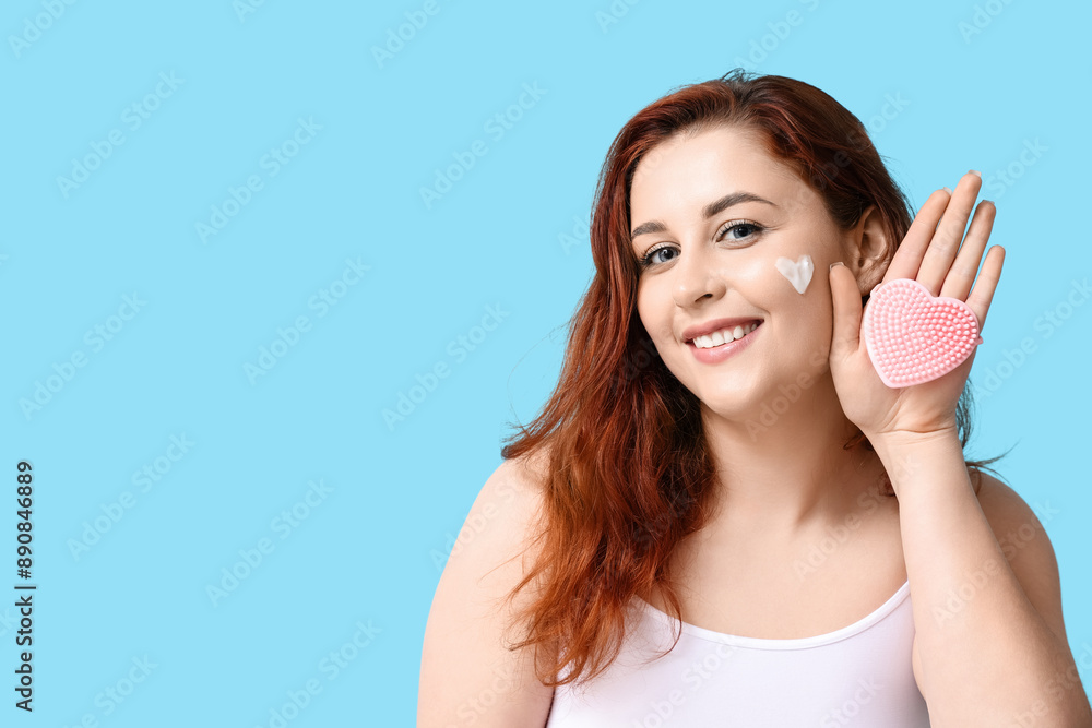 Beautiful young woman with heart-shaped face cream and silicone facial cleansing brush on blue background Beautiful young woman with heart-shaped face cream and silicone facial cleansing brush on blue background
