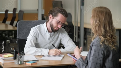 Two professionals sitting on a shared table inside a modern office. Businessman take notes and touch his chin while listening to his workmate explaining about work while holding a mobile phone.