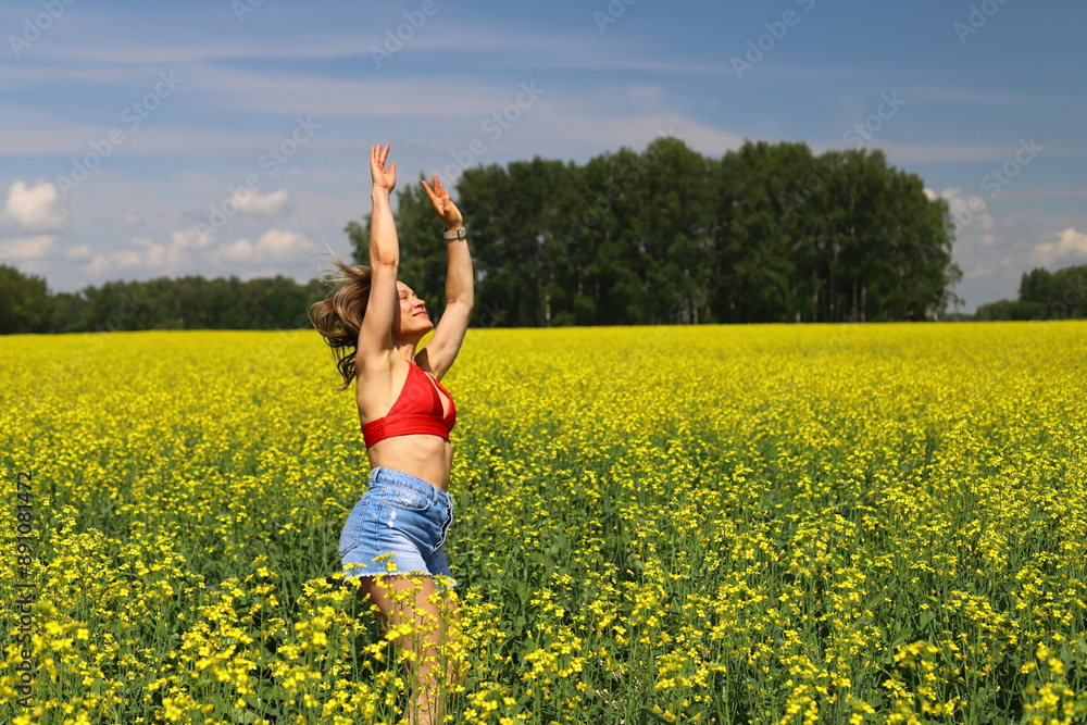 An adult happy woman in a red top and denim shorts jumps and makes a wheel element in a yellow rapeseed field. Motion blur effect. An adult happy woman in a red top and denim shorts jumps and makes a wheel element in a yellow rapeseed field. Motion blur effect.