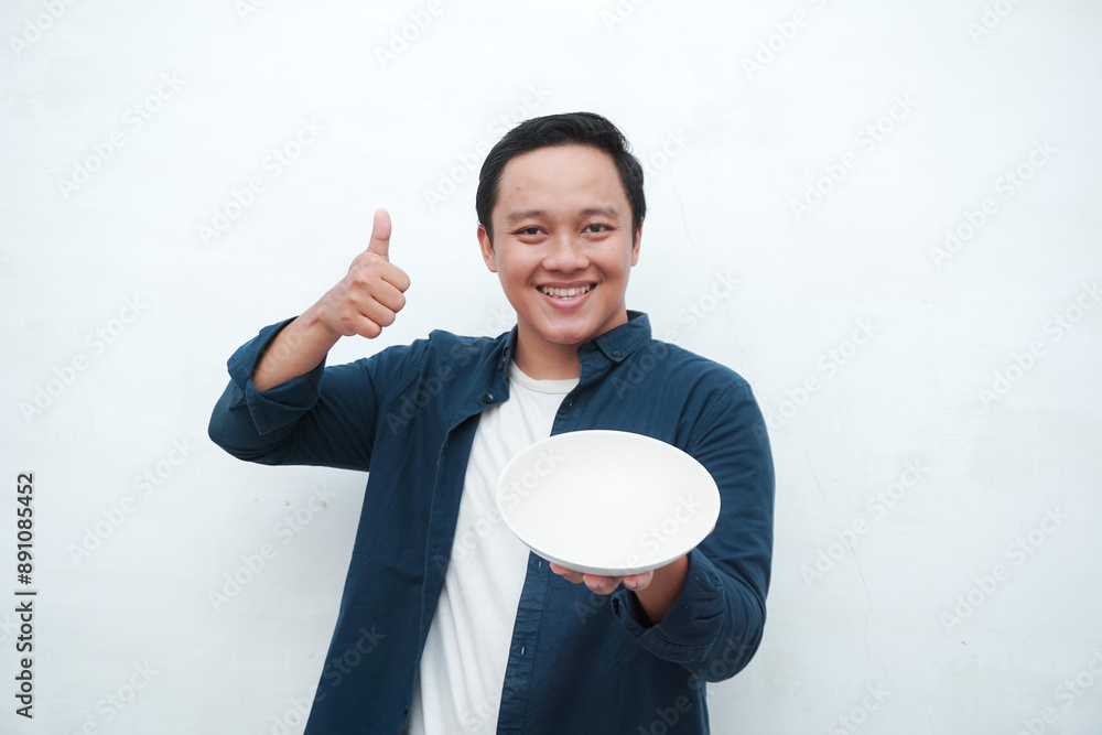Portrait of happy Asian man wearing a blue shirt while holding an empty plate with happy and excited expression. Young man showing and pointing empty mockup plate. Isolated with a white background. Portrait of happy Asian man wearing a blue shirt while holding an empty plate with happy and excited expression. Young man showing and pointing empty mockup plate. Isolated with a white background.