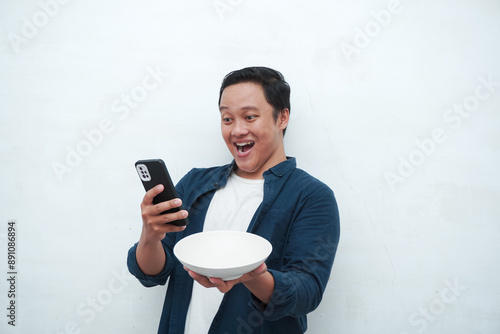 Happy Asian man looking to his phone while holding an empty plate. Concept of ordering food from phone app photo