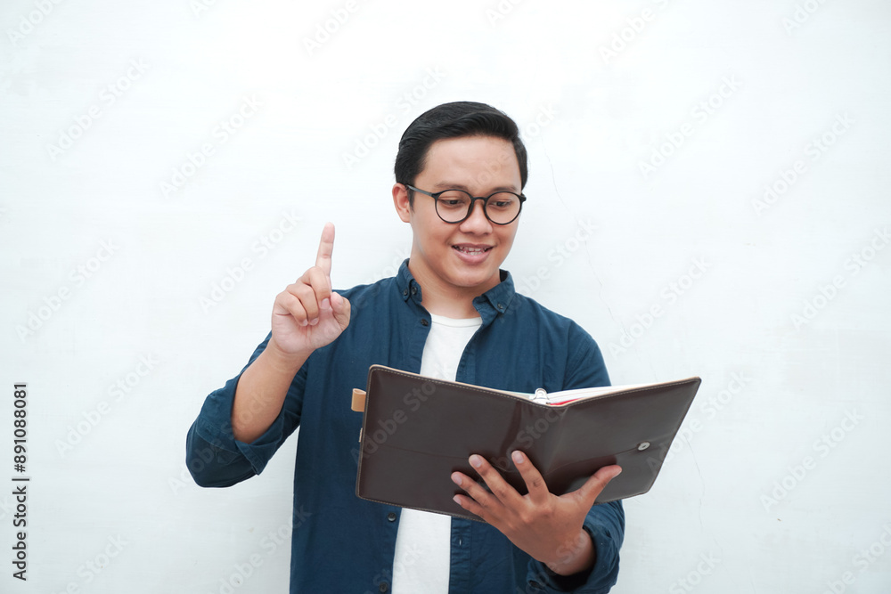 Happy young Asian man with glasses showing expression of gets genius idea. Young Asian man holding books smiling happy and positive. Happy young Asian man with glasses showing expression of gets genius idea. Young Asian man holding books smiling happy and positive.