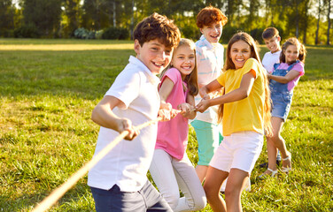Cheerful kids playing fun games on summer day. Group of happy children playing tug of war, standing on green lawn, holding and pulling rope, trying to win victory for their team. Fun, teamwork concept