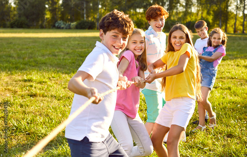 Cheerful kids playing fun games on summer day. Group of happy children playing tug of war, standing on green lawn, holding and pulling rope, trying to win victory for their team. Fun, teamwork concept photo