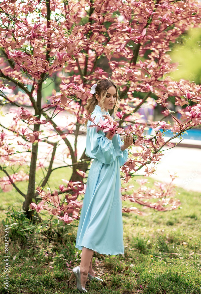 Caucasian woman in blue dress stands by pink magnolia tree Caucasian woman in blue dress stands by pink magnolia tree