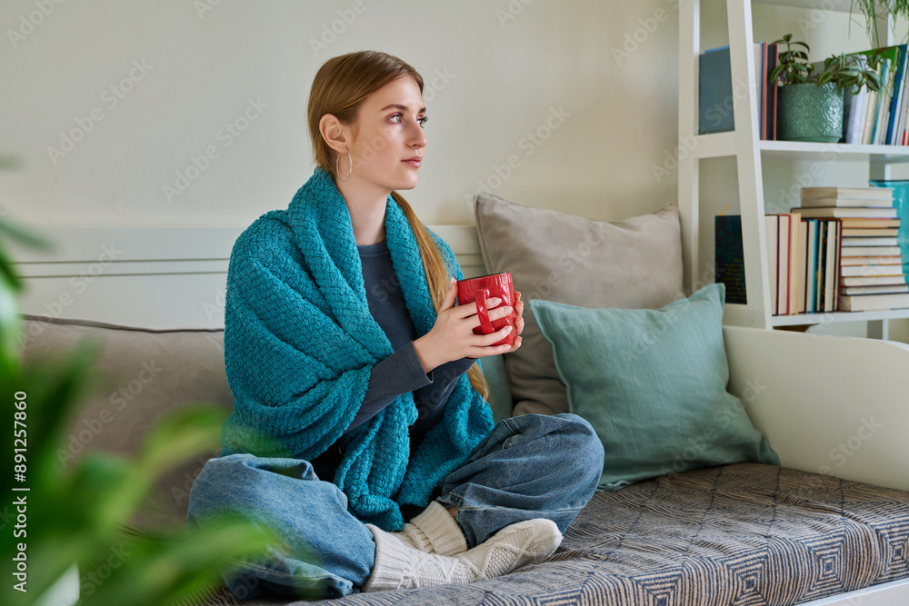 Obraz premium Young woman warming under blanket with mug of hot drink sitting on couch at home