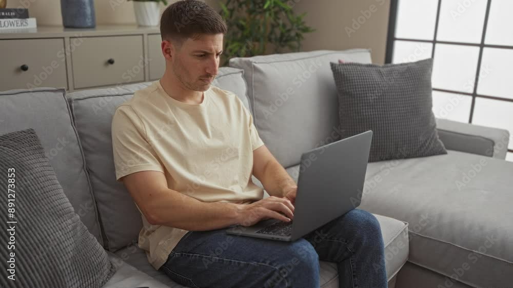 A young man working on a laptop at home in a comfortable living room with a grey couch and modern decor. A young man working on a laptop at home in a comfortable living room with a grey couch and modern decor.