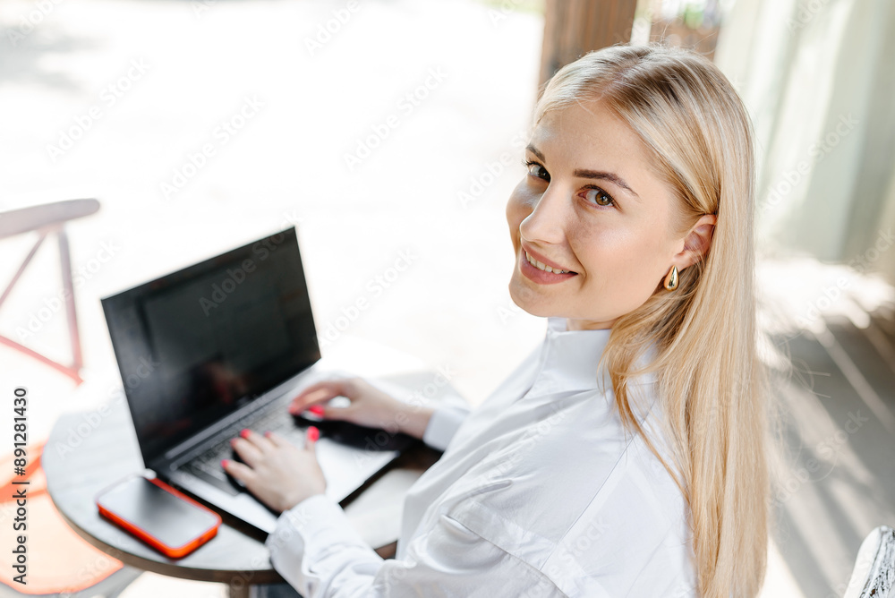 young beautiful blond caucasian woman sitting at table in outdoor terrace of cafe in summer day with notebook, businesswoman working, freelancer typing text and smiling looking at camera young beautiful blond caucasian woman sitting at table in outdoor terrace of cafe in summer day with notebook, businesswoman working, freelancer typing text and smiling looking at camera