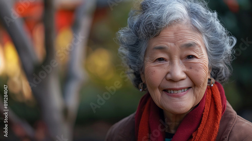 An elderly Asian woman with gray hair, wearing a red scarf, smiling warmly while posing outdoors with a blurred background of greenery.