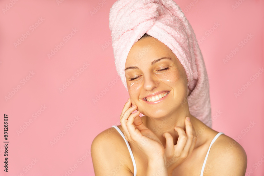 A smiling woman standing in the spa applying eyepatches and a pink towel looking relaxed and happy A smiling woman standing in the spa applying eyepatches and a pink towel looking relaxed and happy
