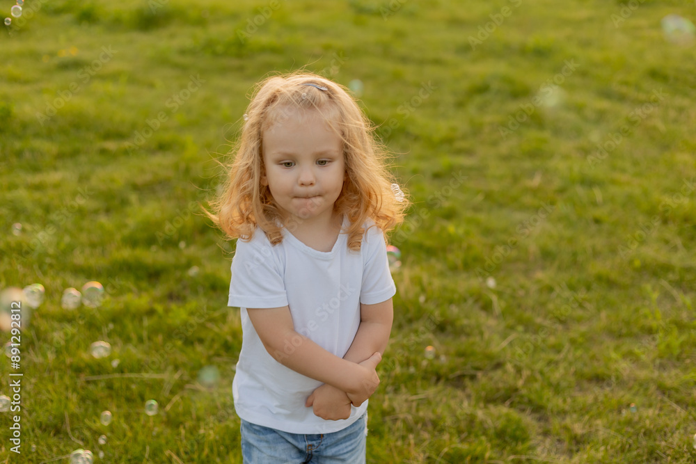 Cute blonde kid with curly hair walks on the lawn with soap bubbles in summer