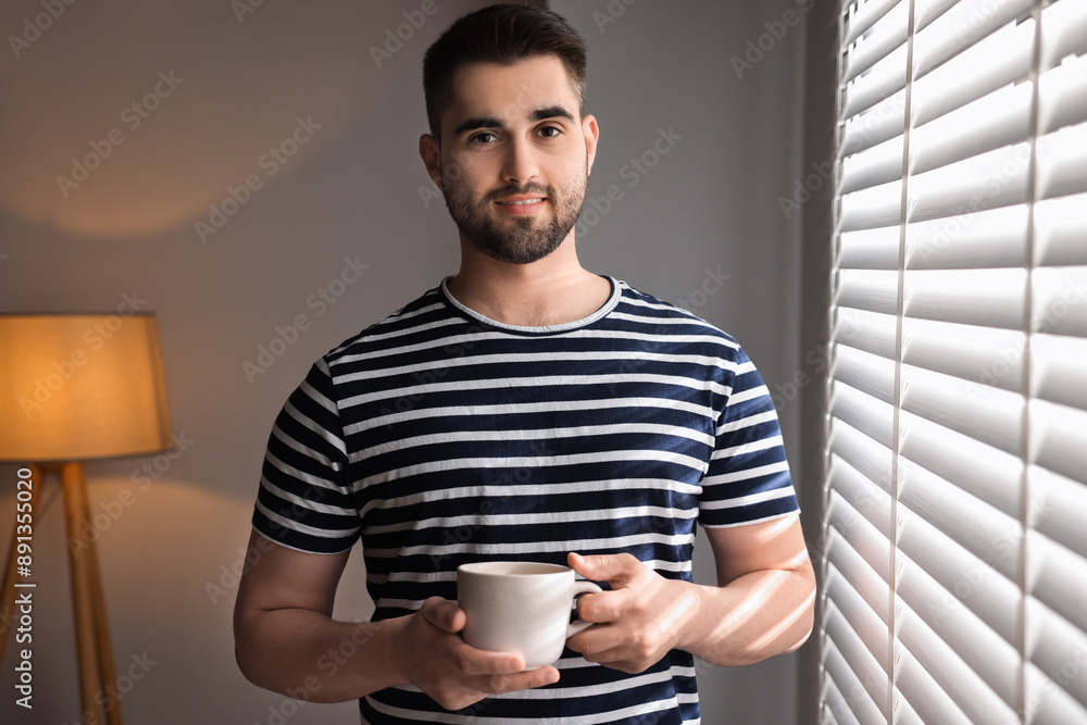 Man with cup of drink near window blinds at home Man with cup of drink near window blinds at home