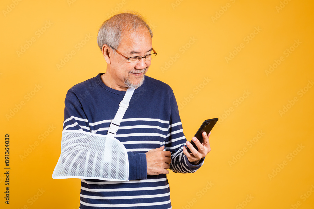 Portrait Asian old man cast his arm and holding mobile phone studio isolated yellow background, retired elderly man accident broken arm wear splint treatment, insurance medical care application Portrait Asian old man cast his arm and holding mobile phone studio isolated yellow background, retired elderly man accident broken arm wear splint treatment, insurance medical care application