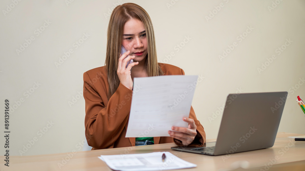 A woman is talking on her cell phone while looking at a piece of paper. She is wearing a brown jacket and is sitting at a desk with a laptop and a cup A woman is talking on her cell phone while looking at a piece of paper. She is wearing a brown jacket and is sitting at a desk with a laptop and a cup