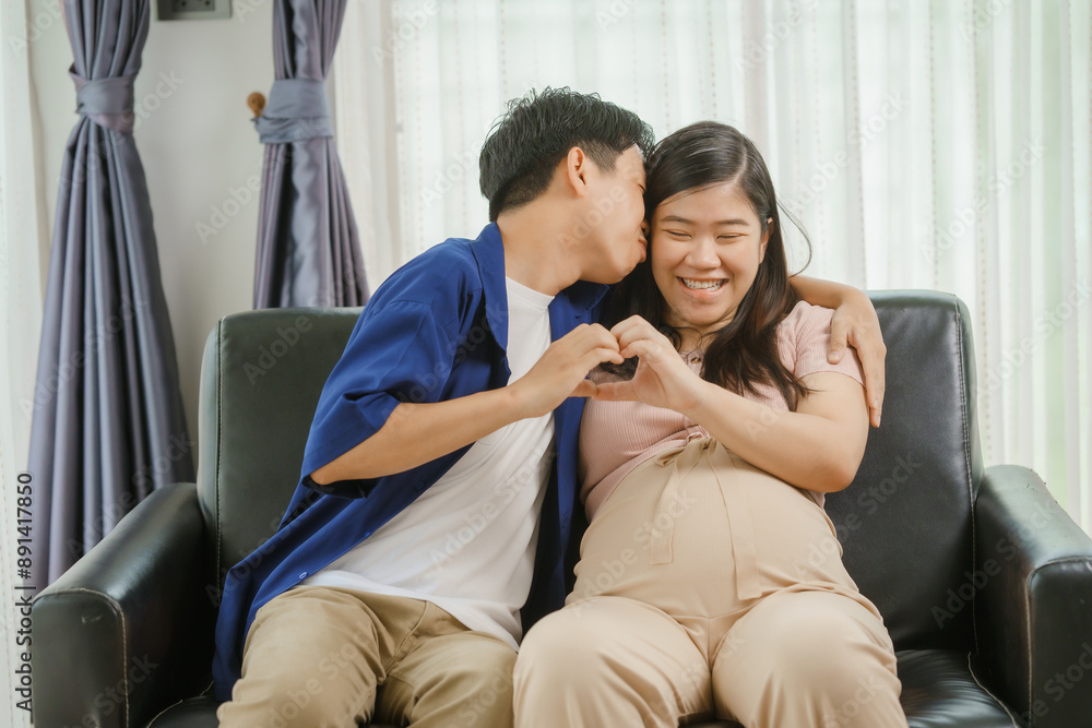 An Asian husband sits on a sofa, gently holding his pregnant wife's stomach. He lovingly cares for her as they eagerly await the arrival of their first child, sharing a tender moment. An Asian husband sits on a sofa, gently holding his pregnant wife's stomach. He lovingly cares for her as they eagerly await the arrival of their first child, sharing a tender moment.
