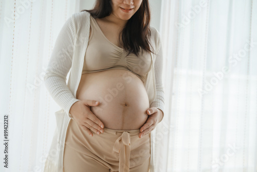 An Asian woman stands while pregnant with her first child, placing her hand on her stomach. She is excited and eagerly waiting for the birth day to meet her baby. Free Download An Asian woman stands while pregnant with her first child, placing her hand on her stomach. She is excited and eagerly waiting for the birth day to meet her baby. - GFXLoad