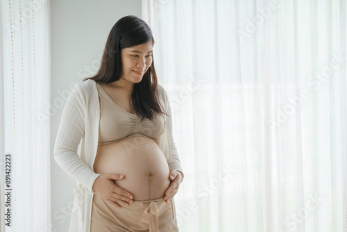 An Asian woman stands while pregnant with her first child, placing her hand on her stomach. She is excited and eagerly waiting for the birth day to meet her baby. Free Download An Asian woman stands while pregnant with her first child, placing her hand on her stomach. She is excited and eagerly waiting for the birth day to meet her baby. - GFXLoad