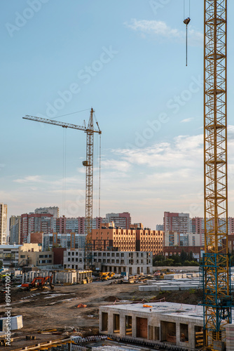 Construction site in the middle of the city in the summer evening. Tower cranes, hydraulic shovel, concrete slabs, construction equipment, vertical photo.
