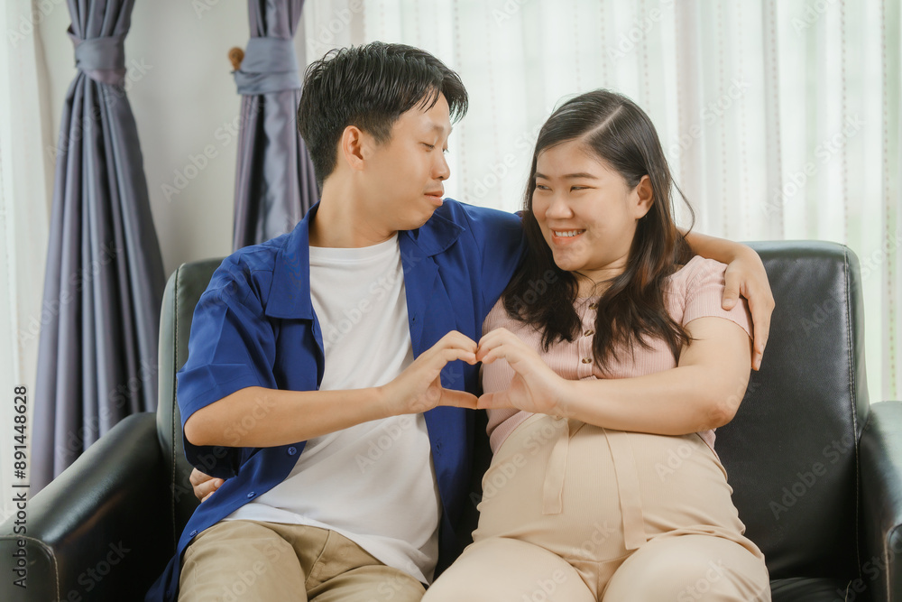 An Asian husband sits on a sofa, gently holding his pregnant wife's stomach. He lovingly cares for her as they eagerly await the arrival of their first child, sharing a tender moment. An Asian husband sits on a sofa, gently holding his pregnant wife's stomach. He lovingly cares for her as they eagerly await the arrival of their first child, sharing a tender moment.