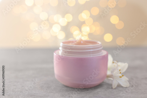 Cream in jar and beautiful jasmine flowers on grey table against light background with blurred lights, closeup