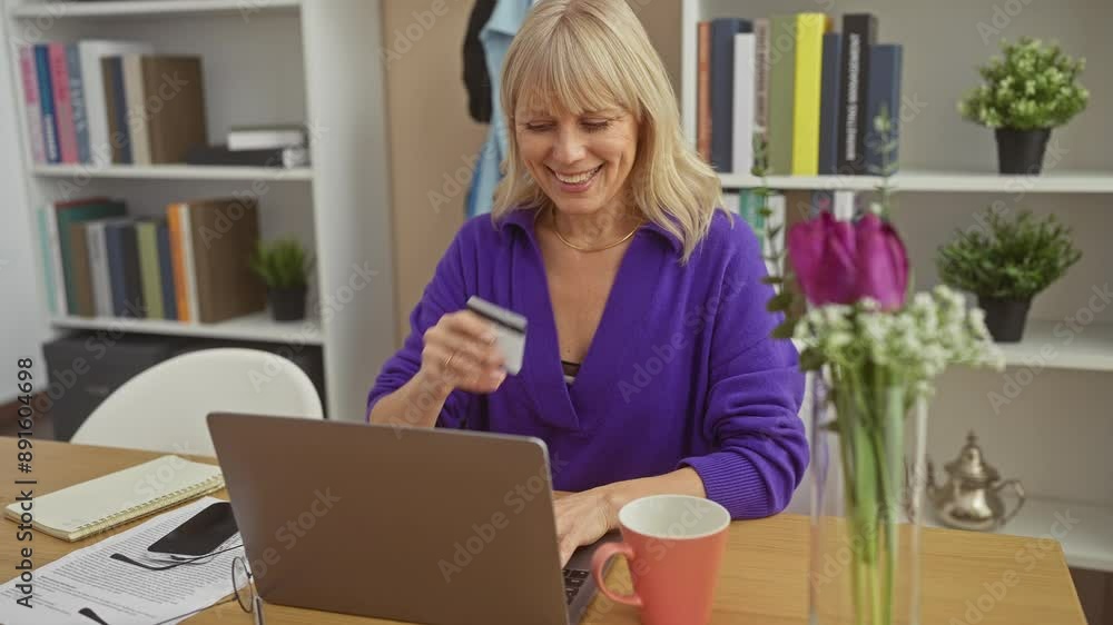 A smiling blonde woman in a purple sweater using a laptop and holding a credit card at a home office. A smiling blonde woman in a purple sweater using a laptop and holding a credit card at a home office.