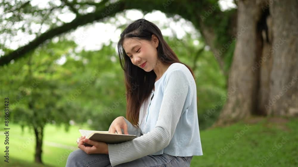 Asian woman sitting on a bench outdoors, reading a notebook. She is wearing a light blue top and jeans, with a coffee cup beside her Asian woman sitting on a bench outdoors, reading a notebook. She is wearing a light blue top and jeans, with a coffee cup beside her