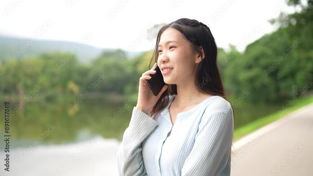 Asian woman talking on a phone while standing on a pathway near a lake. She is wearing a light blue top, with trees in the background. Asian woman talking on a phone while standing on a pathway near a lake. She is wearing a light blue top, with trees in the background.