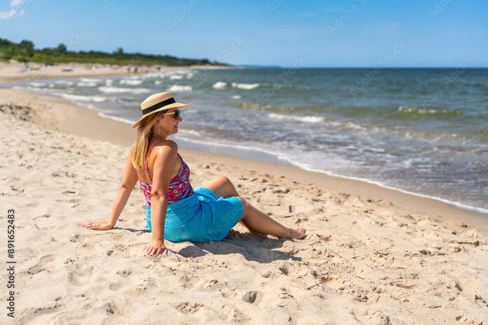 Summer holidays on the Baltic Sea. Beautiful young woman in a colorful swimsuit and a blue pareo and hat sitting on the ground on a sandy beach by the sea, looking dreamily at the horizon. Side view Summer holidays on the Baltic Sea. Beautiful young woman in a colorful swimsuit and a blue pareo and hat sitting on the ground on a sandy beach by the sea, looking dreamily at the horizon. Side view