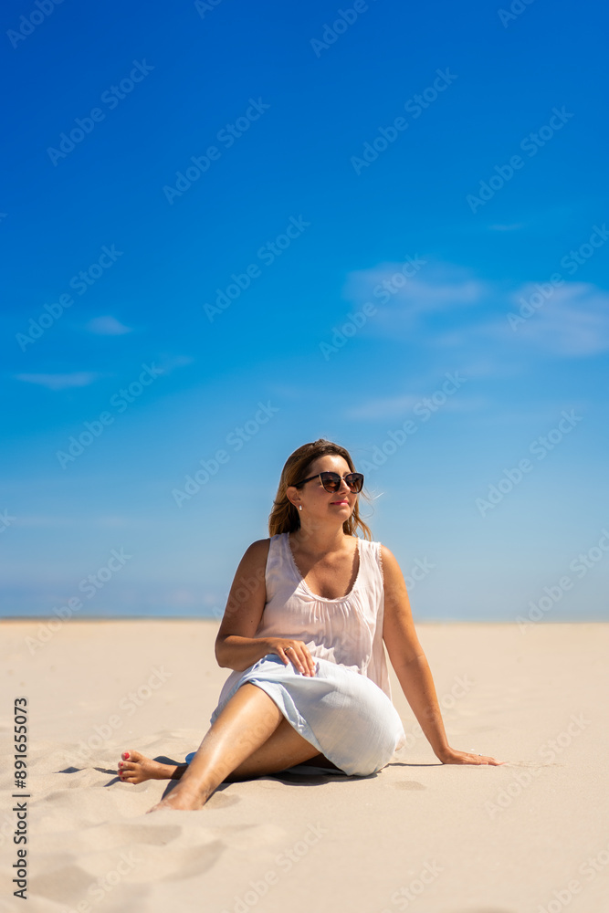 Summer holidays. Portrait of young beautiful woman with long blonde hair in light dress and sunglasses sitting on ground on sandy beach on sunny day. Energy, joy of life and youth Summer holidays. Portrait of young beautiful woman with long blonde hair in light dress and sunglasses sitting on ground on sandy beach on sunny day. Energy, joy of life and youth