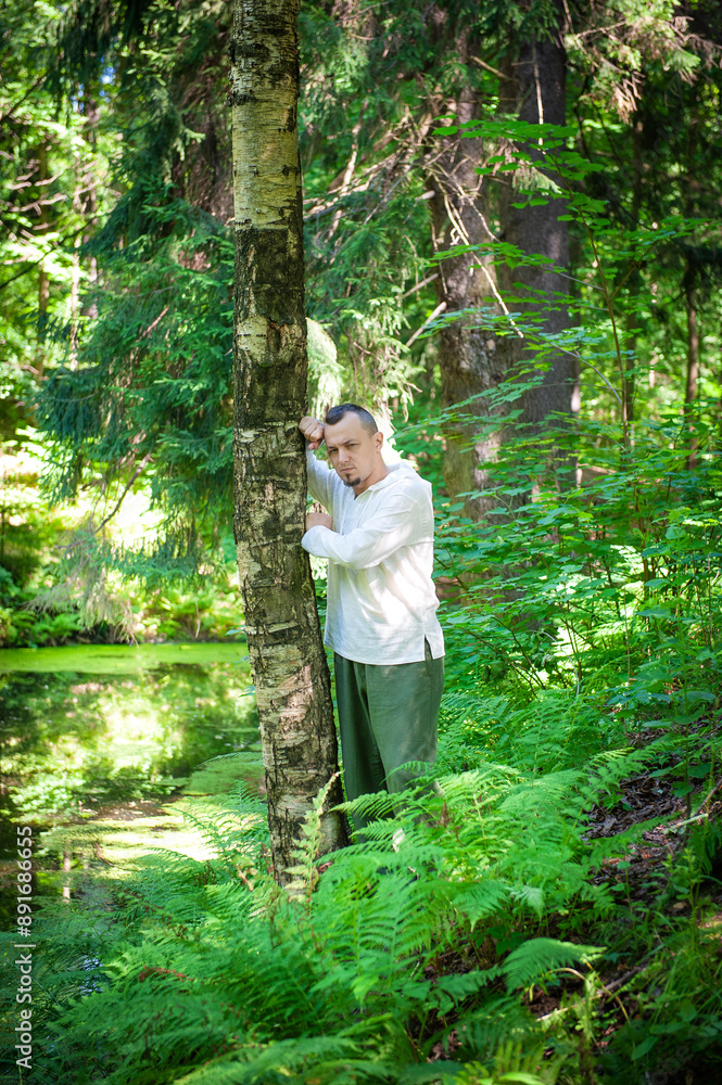 Young sad man in traditional Slavic clothes stand near a tree in the forest. Thoughtful and pensive, he reflects on solitude Young sad man in traditional Slavic clothes stand near a tree in the forest. Thoughtful and pensive, he reflects on solitude