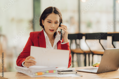 Businesswoman on the Phone: A focused businesswoman in a red blazer, multitasks by reviewing documents while talking on the phone, showcasing the hustle of modern business. 