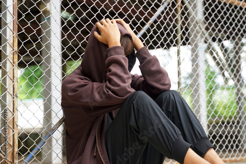 Canvas Print A young Asian male wearing a hood sits with his face in his hands and his head in his hands, his back resting against a wire mesh fence in a juvenile detention center