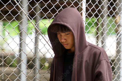 Thai male teenager wearing a hoodie Covering his face, he sat sadly alone with his back against the steel wall of the detention center.