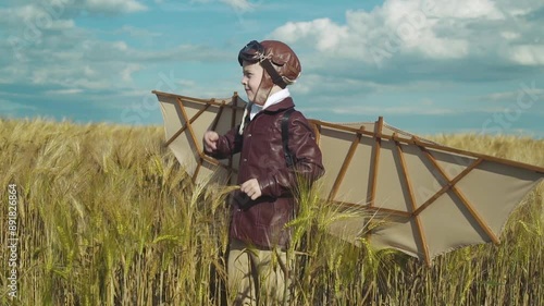 Little boy in pilot suit with artificial wings on his back stands in wheat field