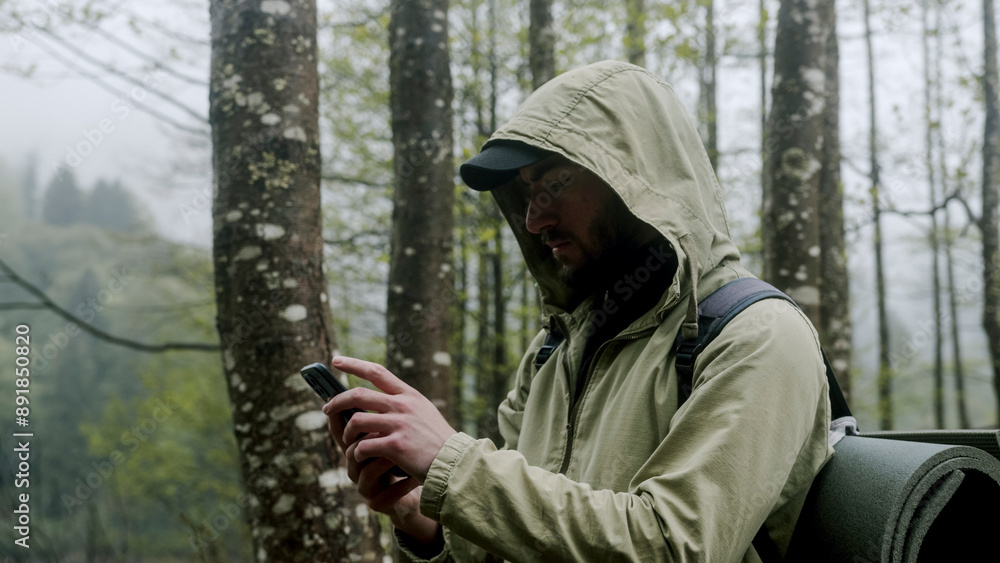 A hiker in a forest using a smartphone, wearing a hooded jacket and a backpack. The hiker is checking his phone, possibly for navigation or communication, in a misty and wooded environment A hiker in a forest using a smartphone, wearing a hooded jacket and a backpack. The hiker is checking his phone, possibly for navigation or communication, in a misty and wooded environment
